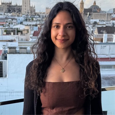 Rhea, a woman with long curly dark hair wearing a brown top and gold necklace, smiling on a balcony with a city skyline behind her.